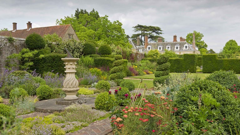 A view of Woolbeding Gardens with a sundial to the left, surrounded by topiary hedges and beds of flowers